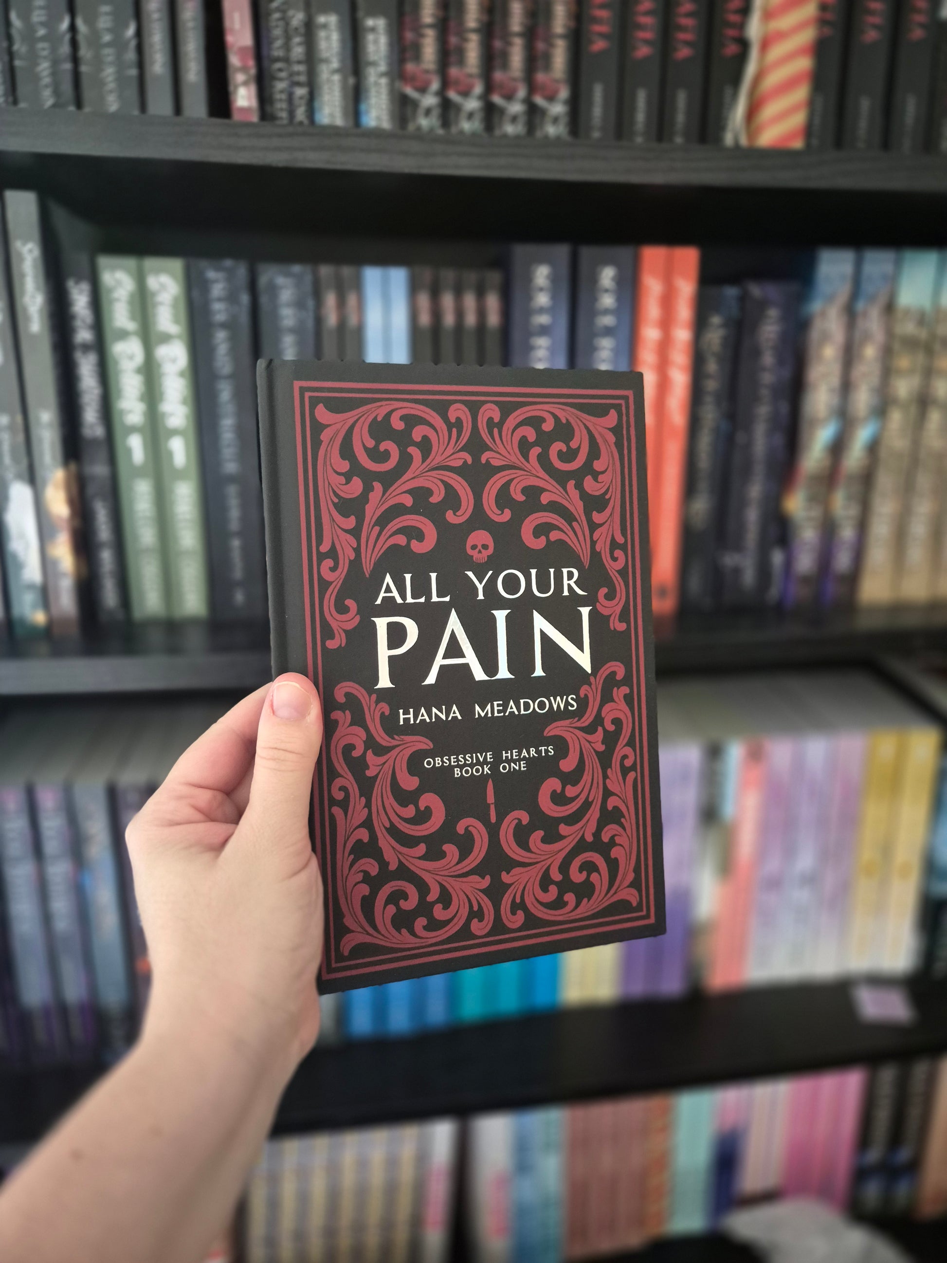 Hand holding a book titled 'All Your Pain' in front of a bookshelf.