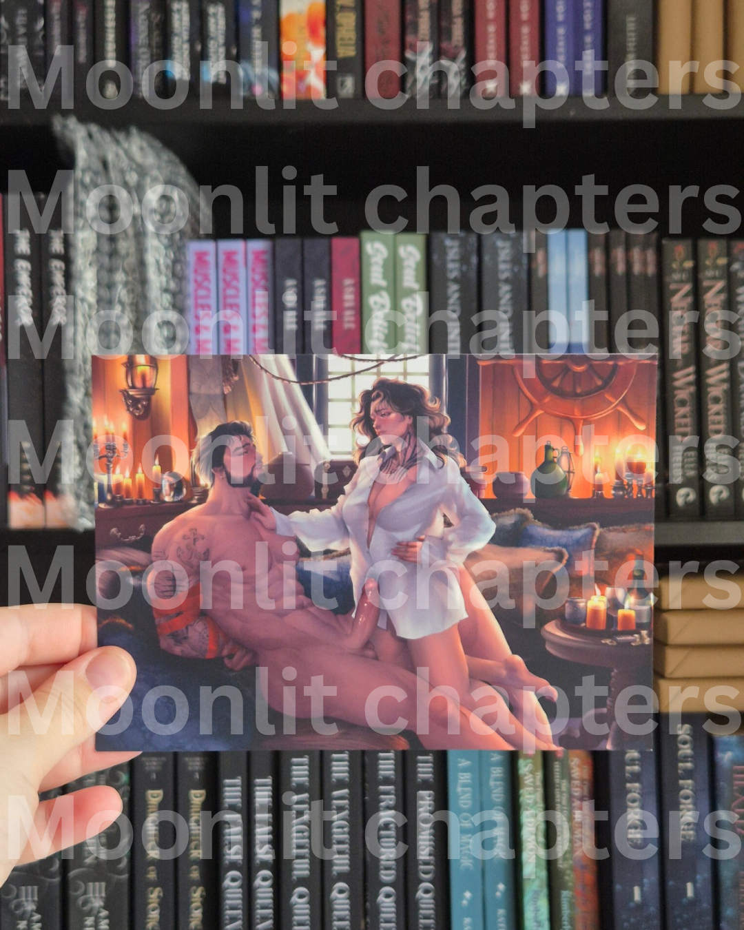 A hand holds a printed erotic art card featuring a man and woman in a warmly lit room with candles, in front of a bookshelf. A large repeated watermark reading 'Moonlit chapters' overlays the entire photo.