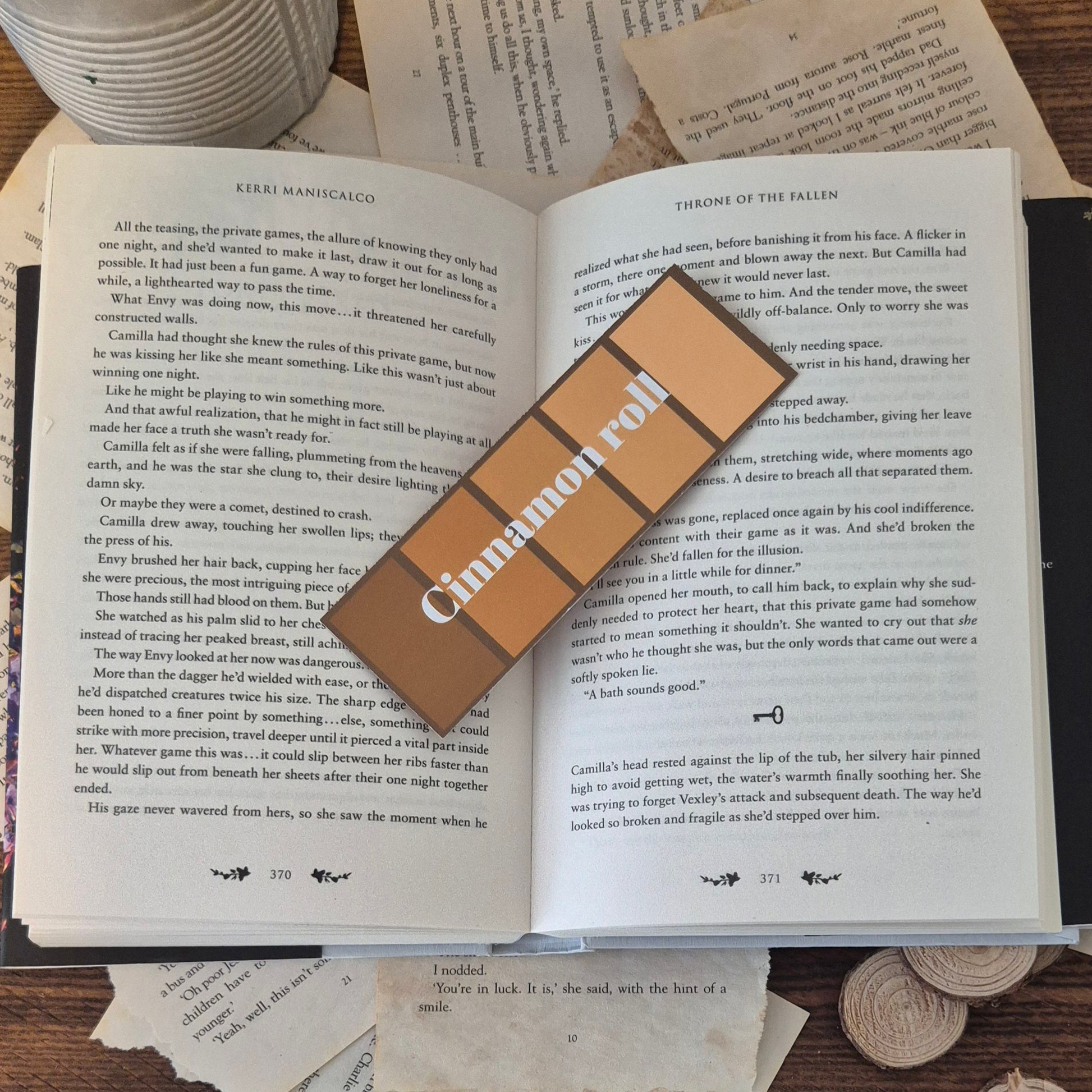 The cinnamon roll bookmark is pictured ontop an open book. It is also surrpunded by a fake plant, and a collection of ripped and aged book pages 