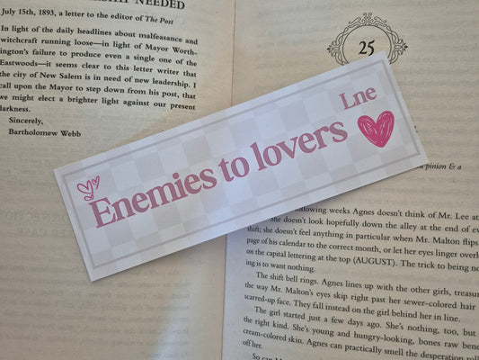 Bookmark with a chequered background with the words 'enemies to lovers Lne' on a open book