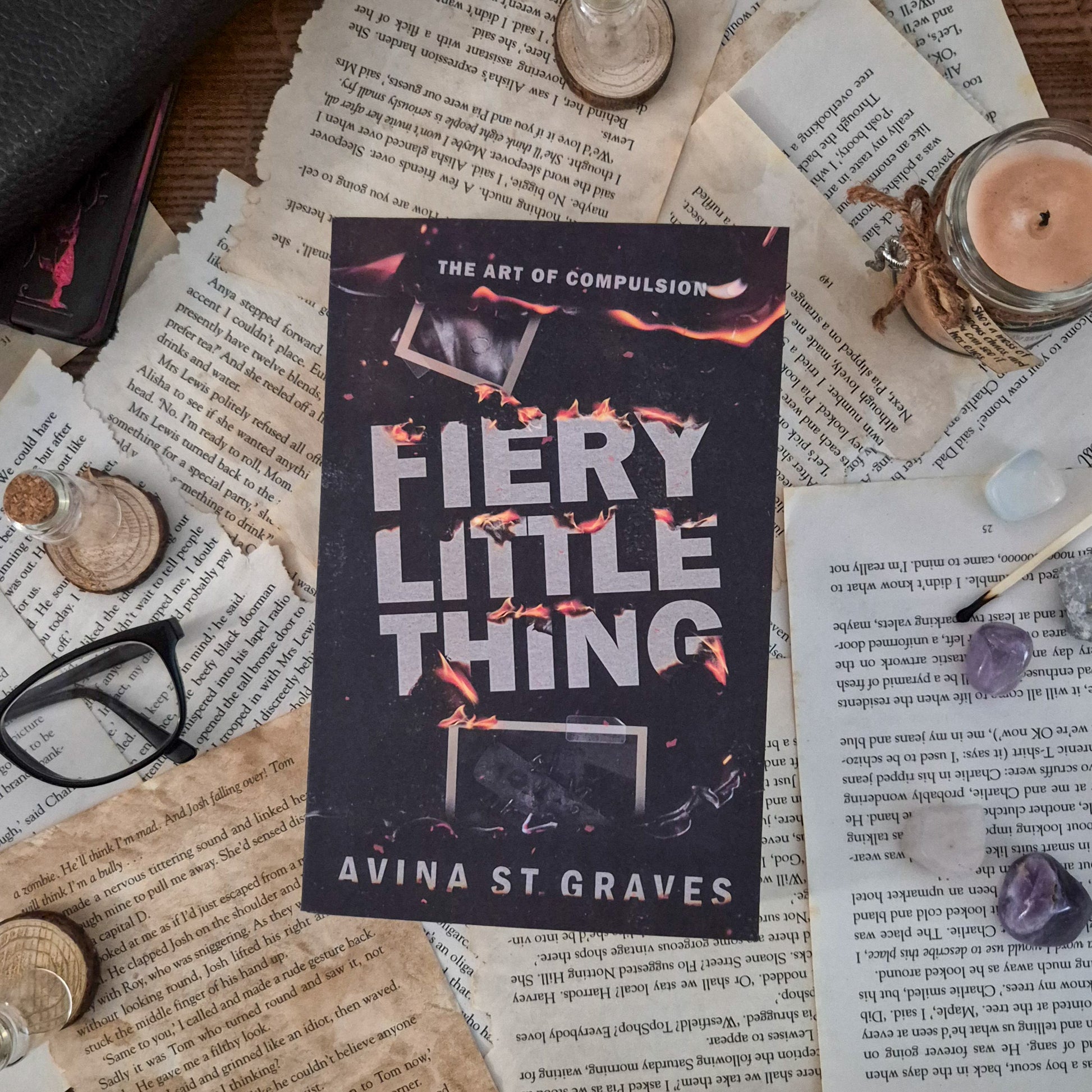 Fiery little thing by Avina St Graves. A dark romance. The product can be seen on a bed of ripped, aged pages. Surrounded by objects such as a pair of glassss, a stack of notebooks, candles and a used matchstick
