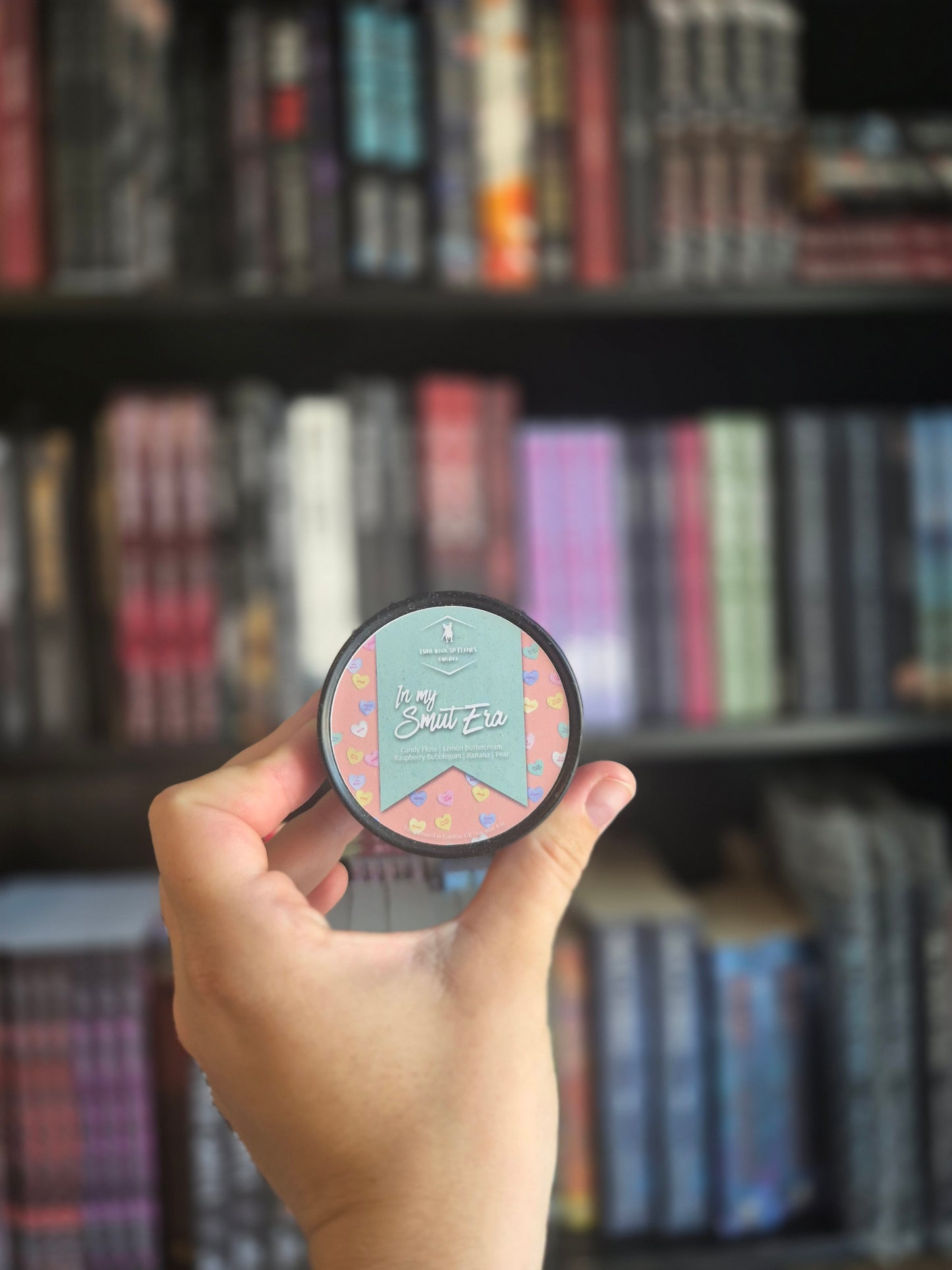 A hand holding a round bookish candle with a light blue and pink patterned wax design, against a blurred background of shelves filled with books