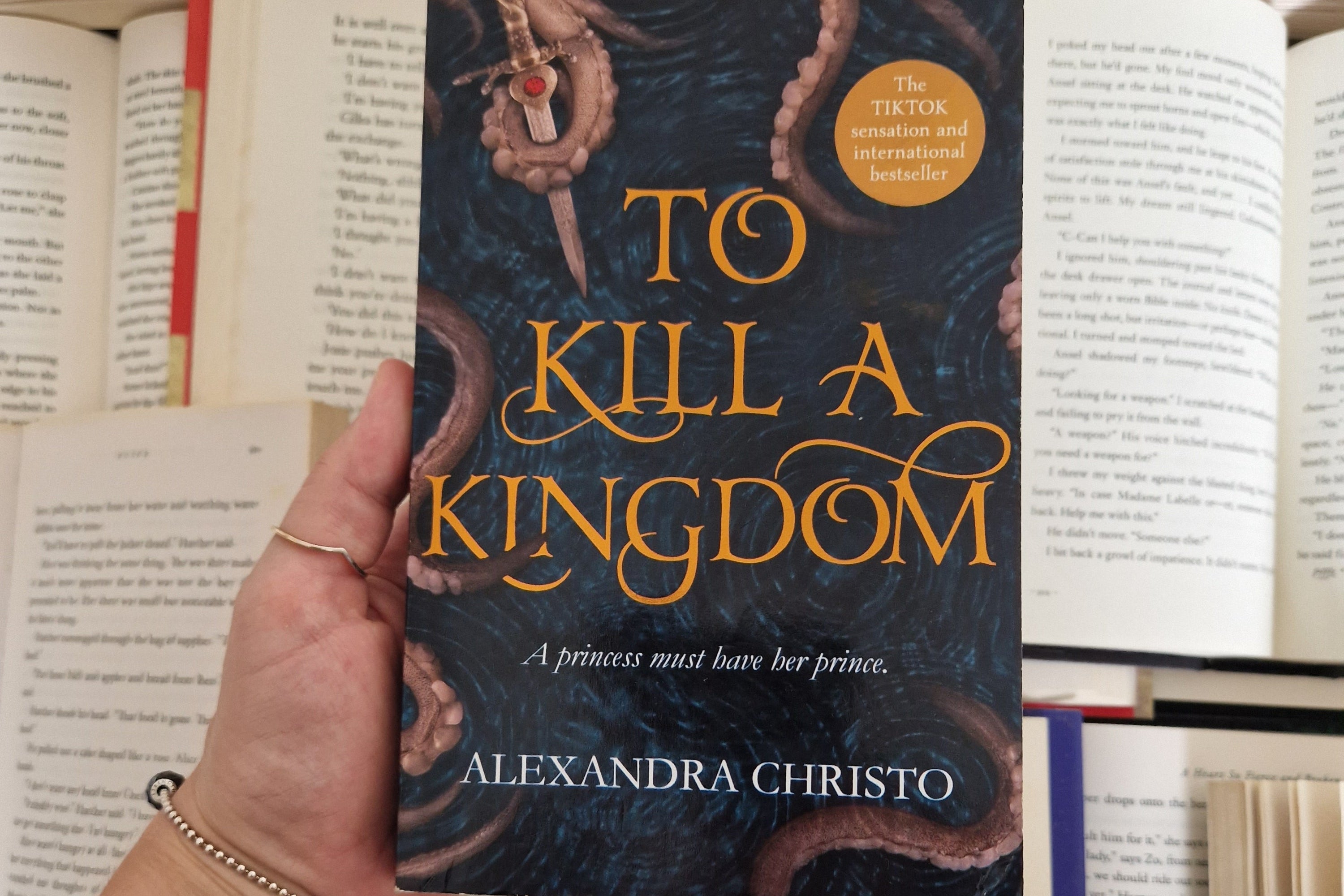 To kill a kingdom by Alexandra Christa, held over a collection of open hardback books.
